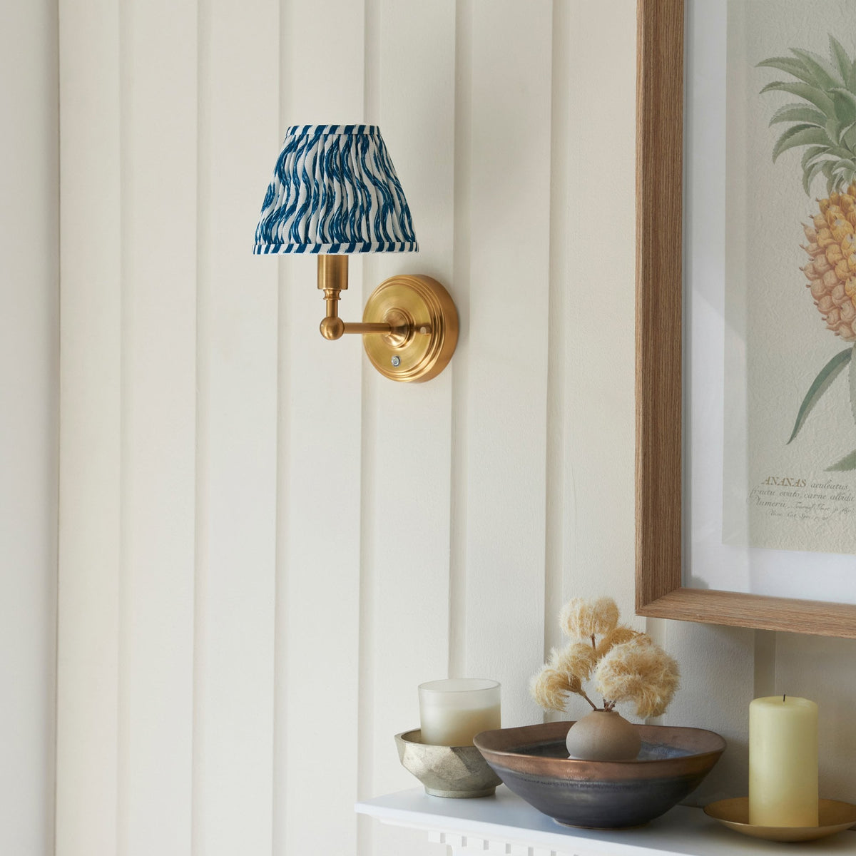 A brass wall light with a blue and white patterned pleated shade on a paneled wall.