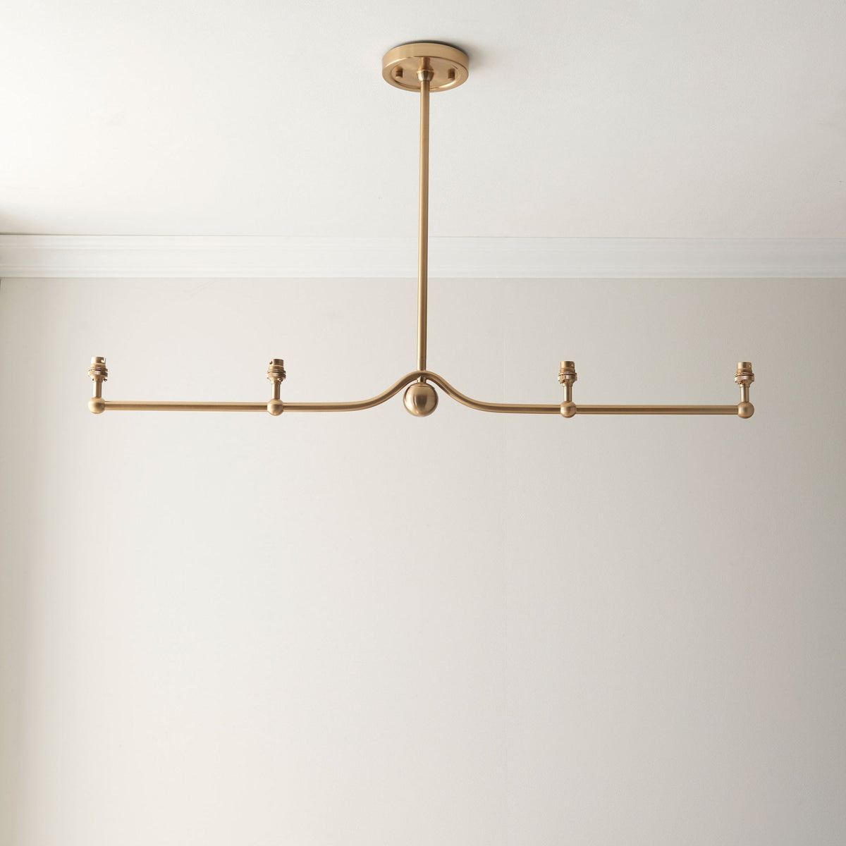 A horizontal four-light gold pendant fitting without shades against a white wall.
