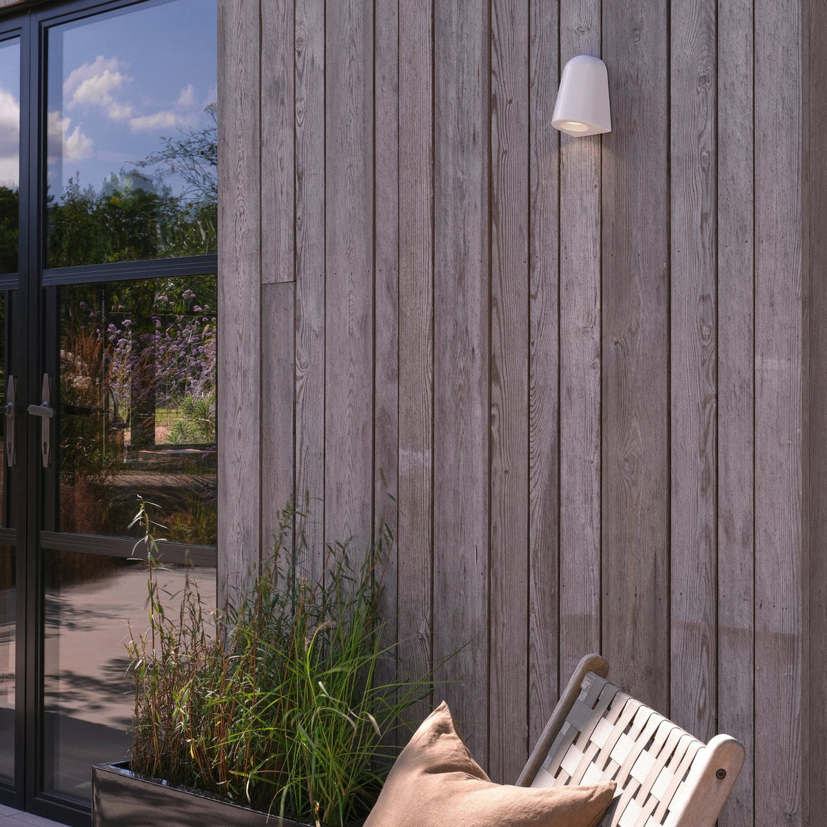 White outdoor wall light on a wooden wall, with a chair and plant pot in the foreground