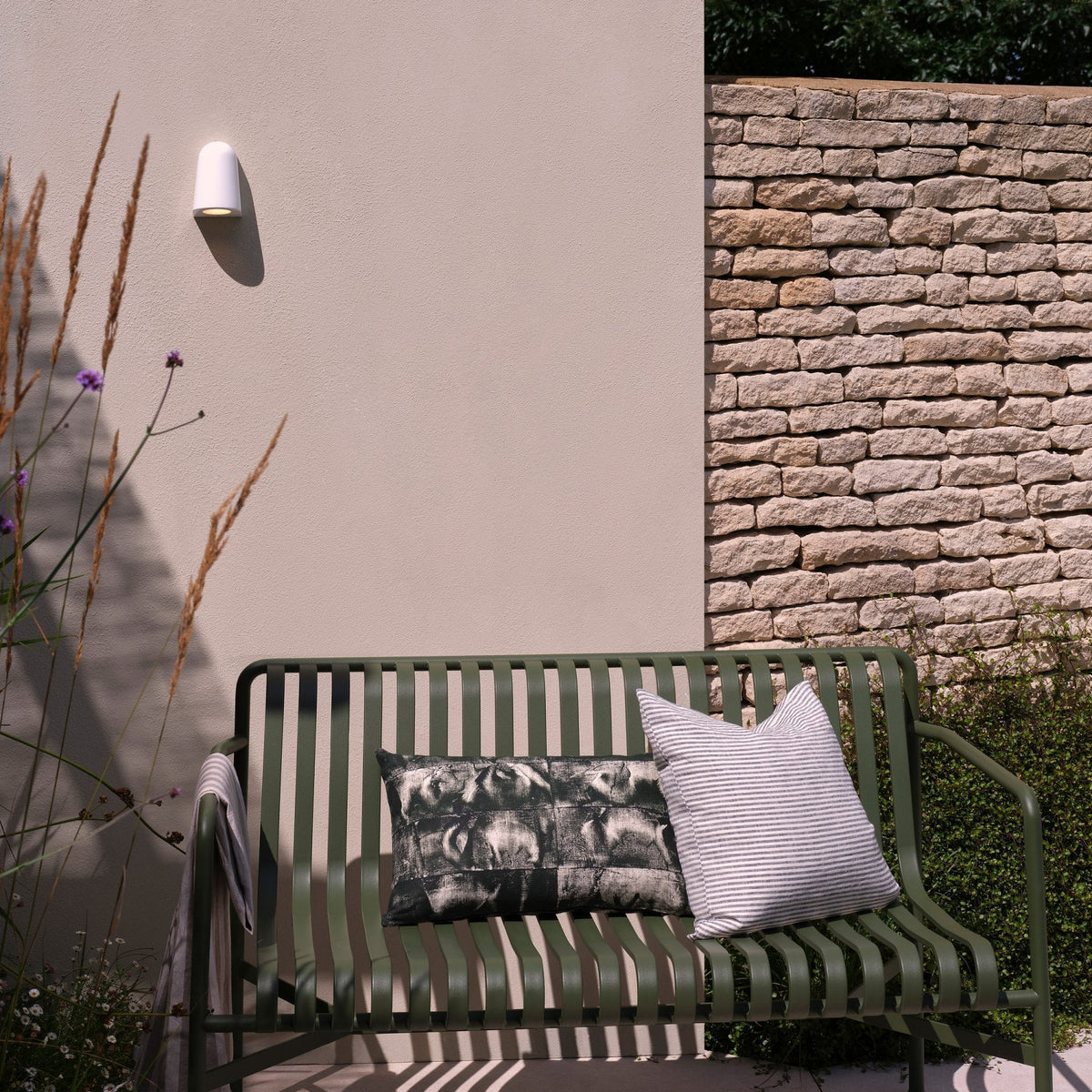 Green metal garden bench with two cushions, next to a light pink wall with a white outdoor light