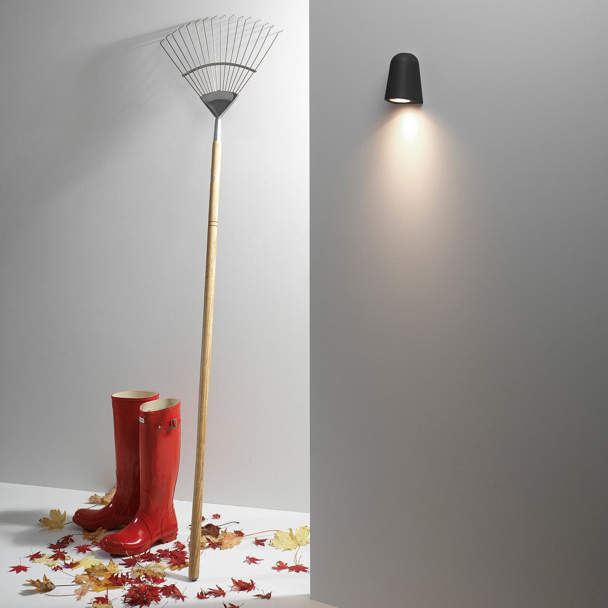 Black outdoor wall light illuminates a wall next to a rake, red boots, and autumn leaves