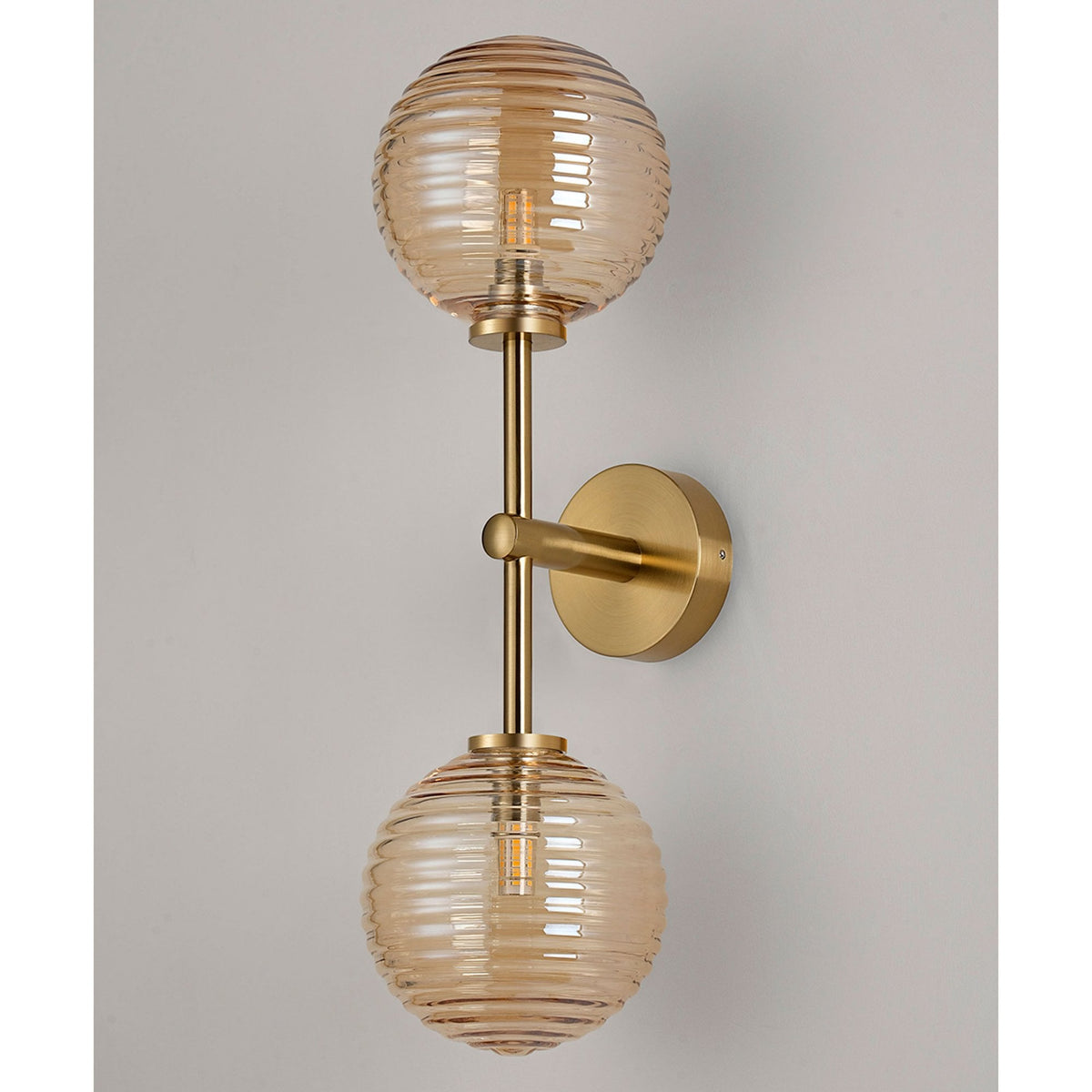 A brass wall light with two amber ribbed glass globe shades against a light grey wall.