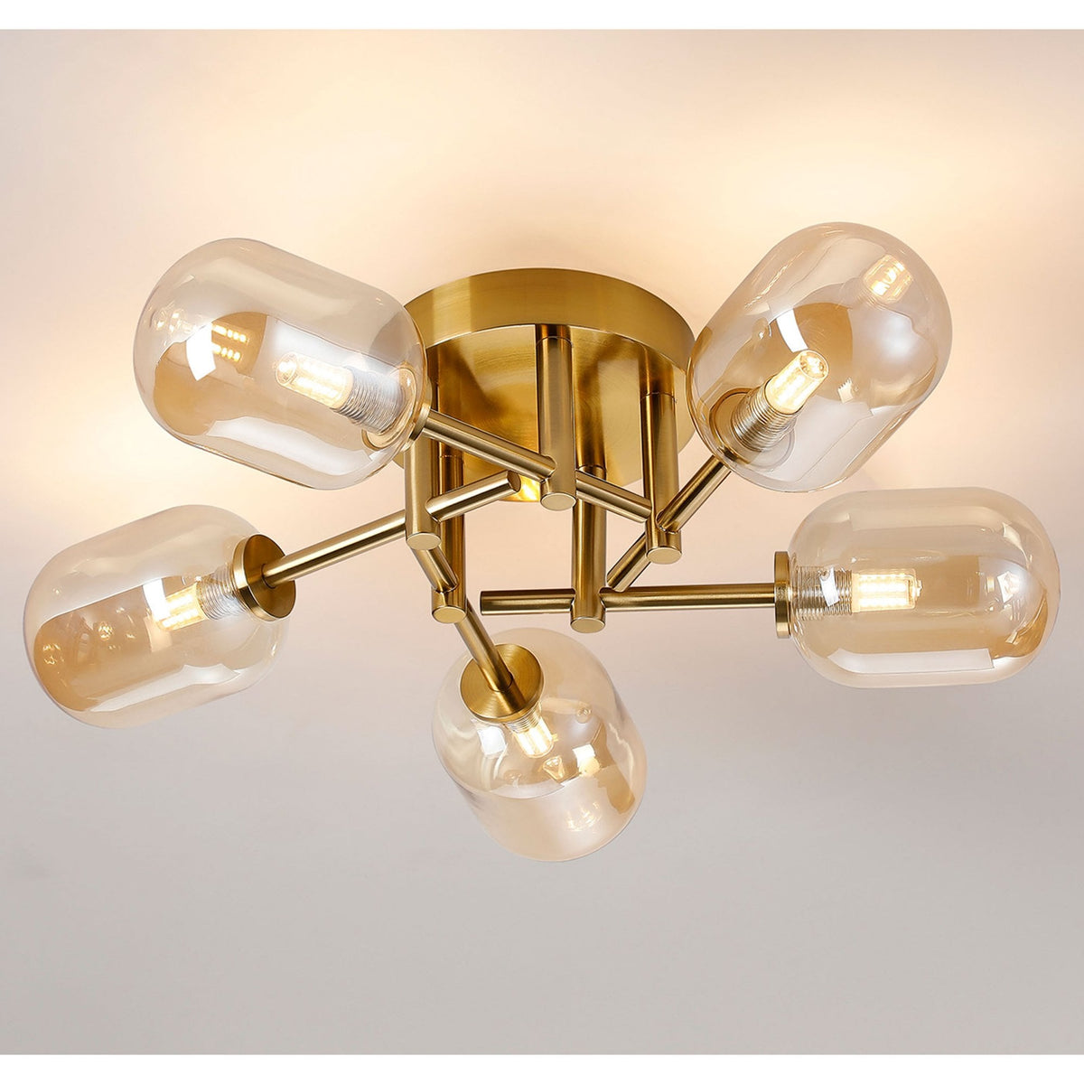 A modern brass ceiling light with five amber glass shades, illuminated against a white ceiling.
