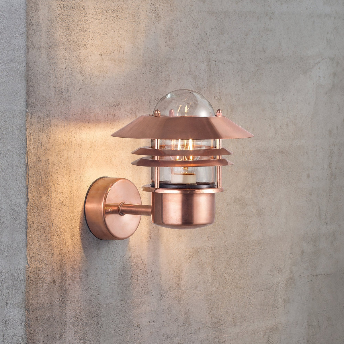 A copper outdoor wall light with tiered shades illuminates a concrete wall at dusk.