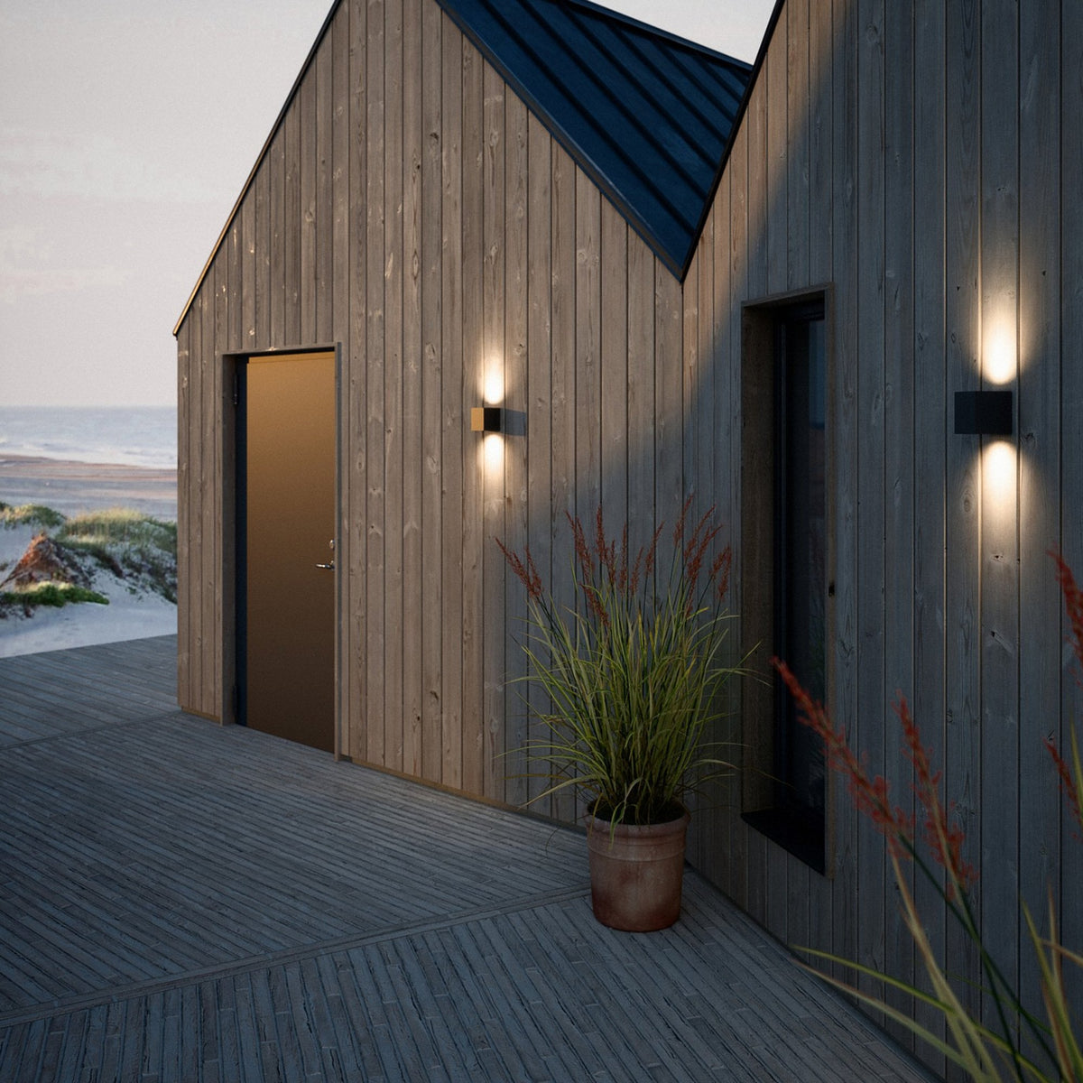 Two exterior wall lights illuminating a wooden building and decking at dusk by the sea.