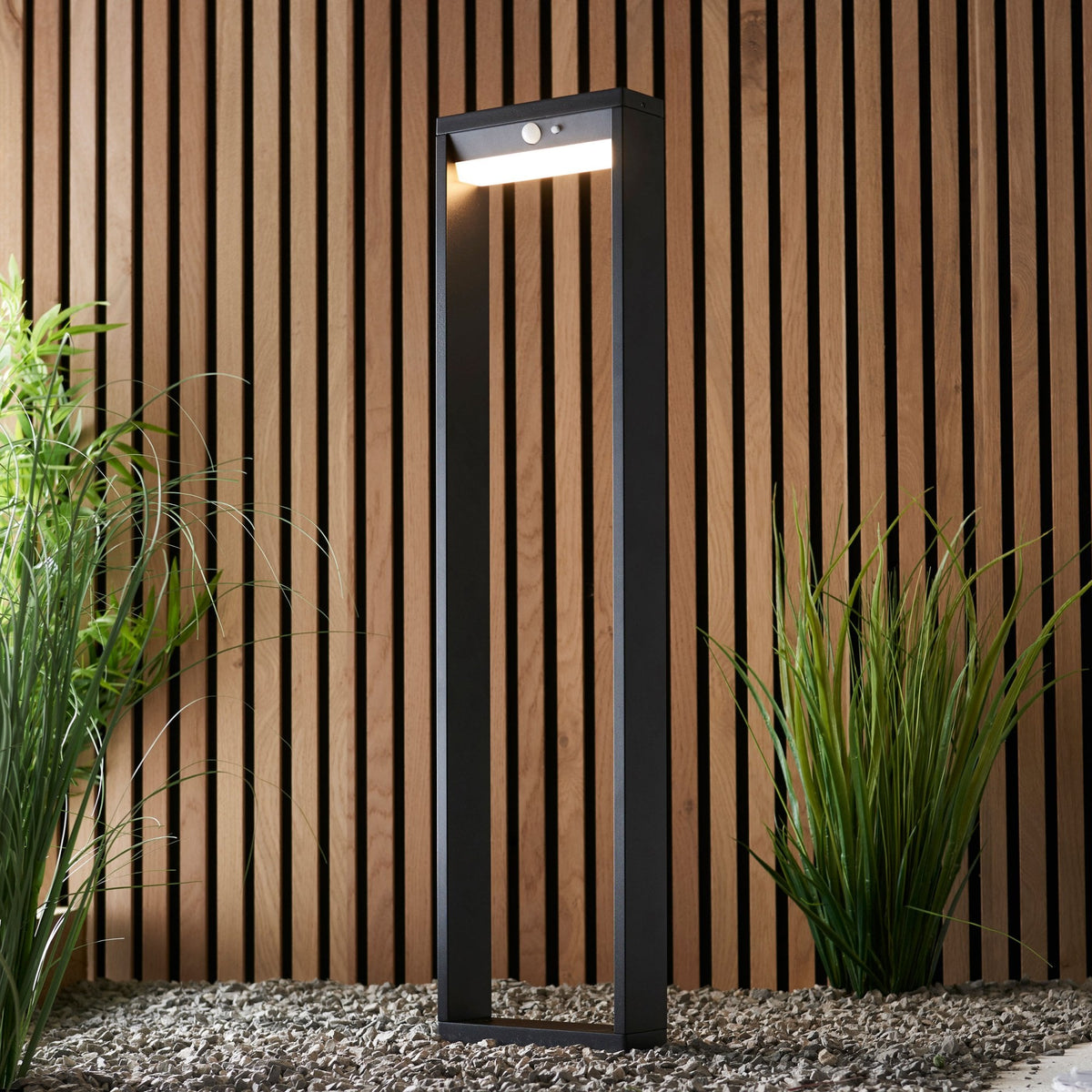 Black outdoor bollard light with a motion sensor, illuminating a garden path with a wooden slatted fence.
