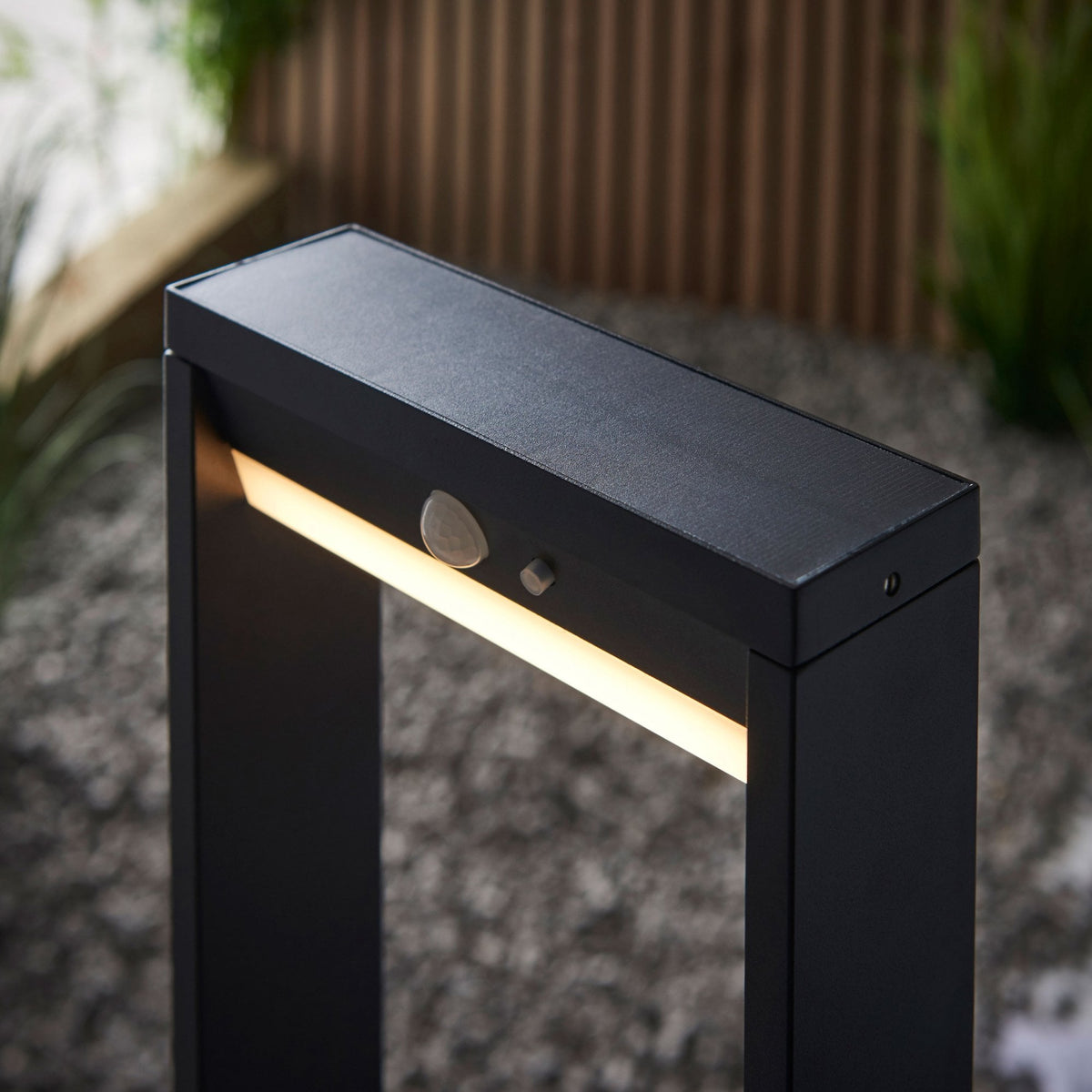 Top-down view of a black outdoor bollard light with a motion sensor, set in a gravel garden.
