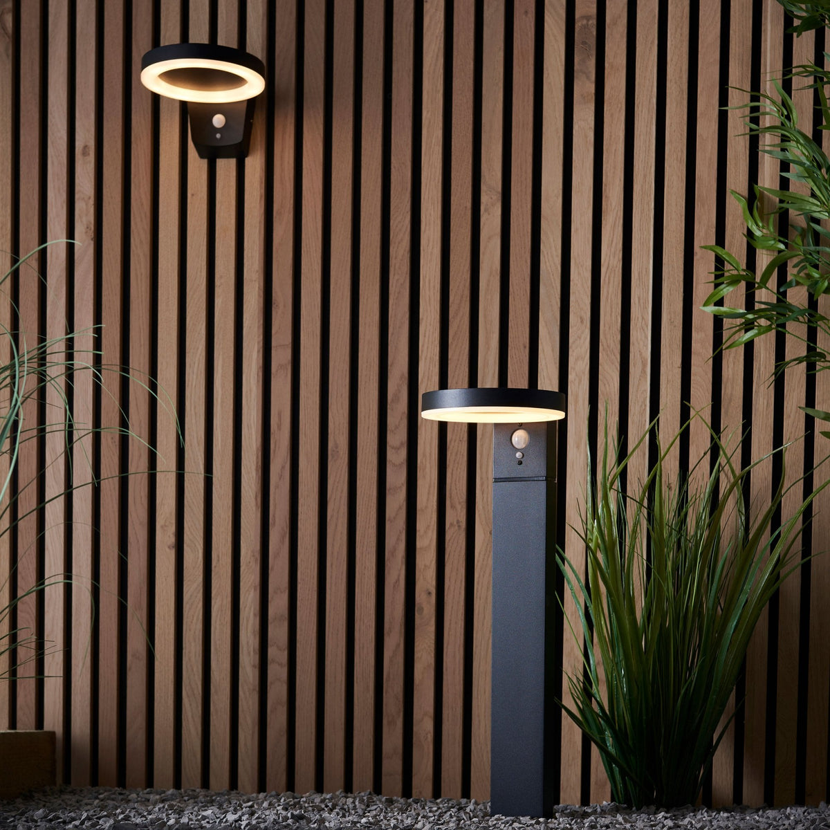 A black bollard light and a matching wall light illuminate a garden with a wooden fence.
