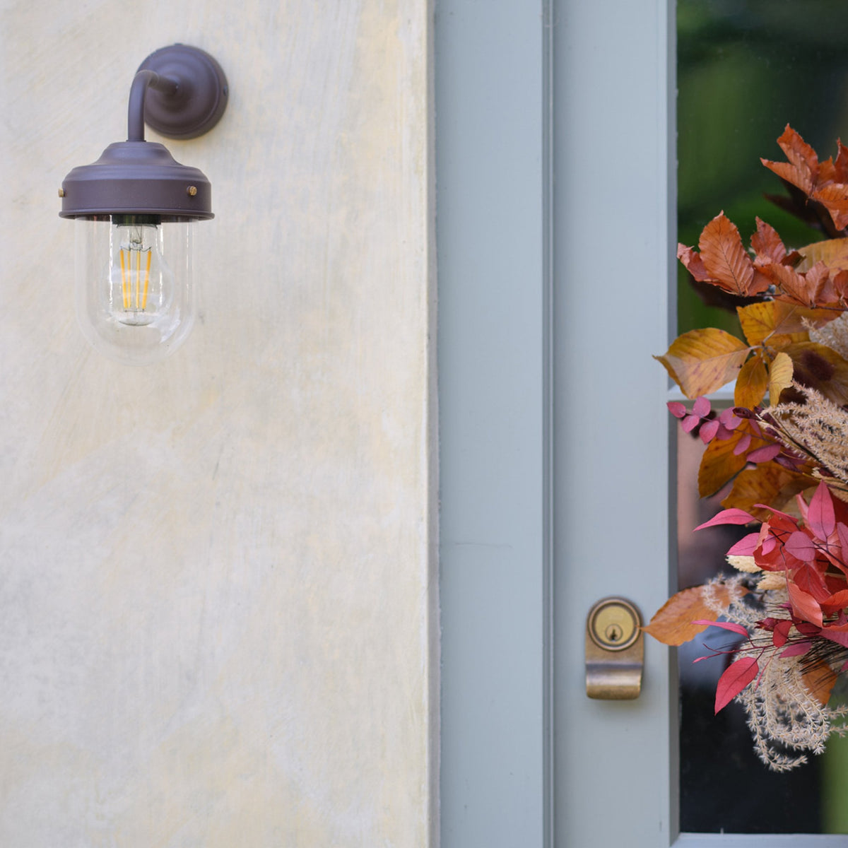 A bronze outdoor wall light with a clear glass shade and an Edison bulb, next to a door with autumn decor.