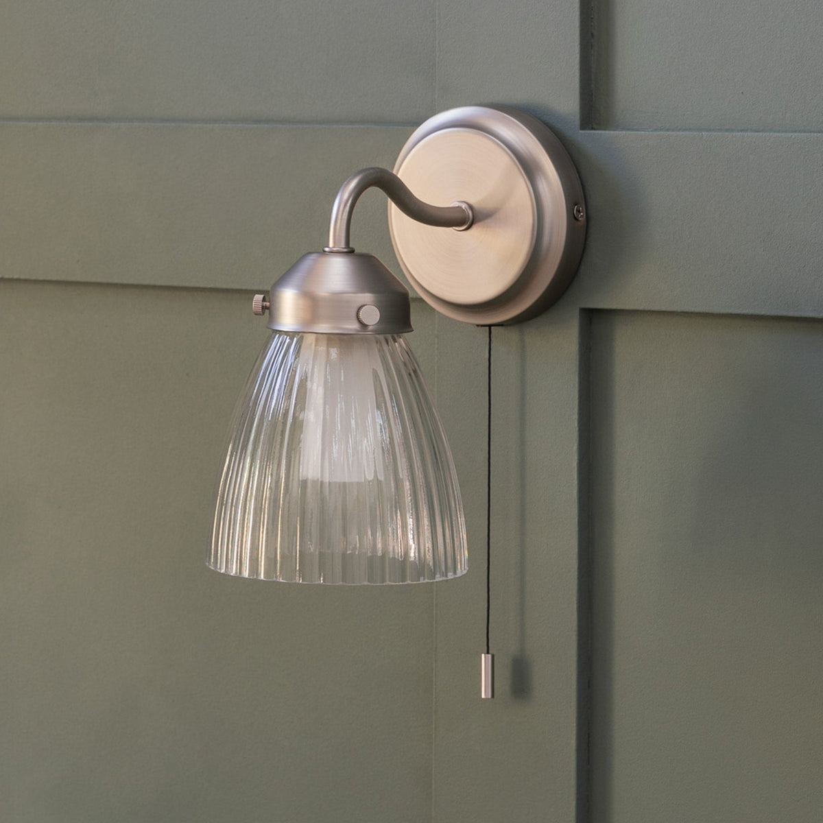 Brushed chrome wall light with a ribbed glass shade and a pull cord switch, mounted on a green paneled wall.