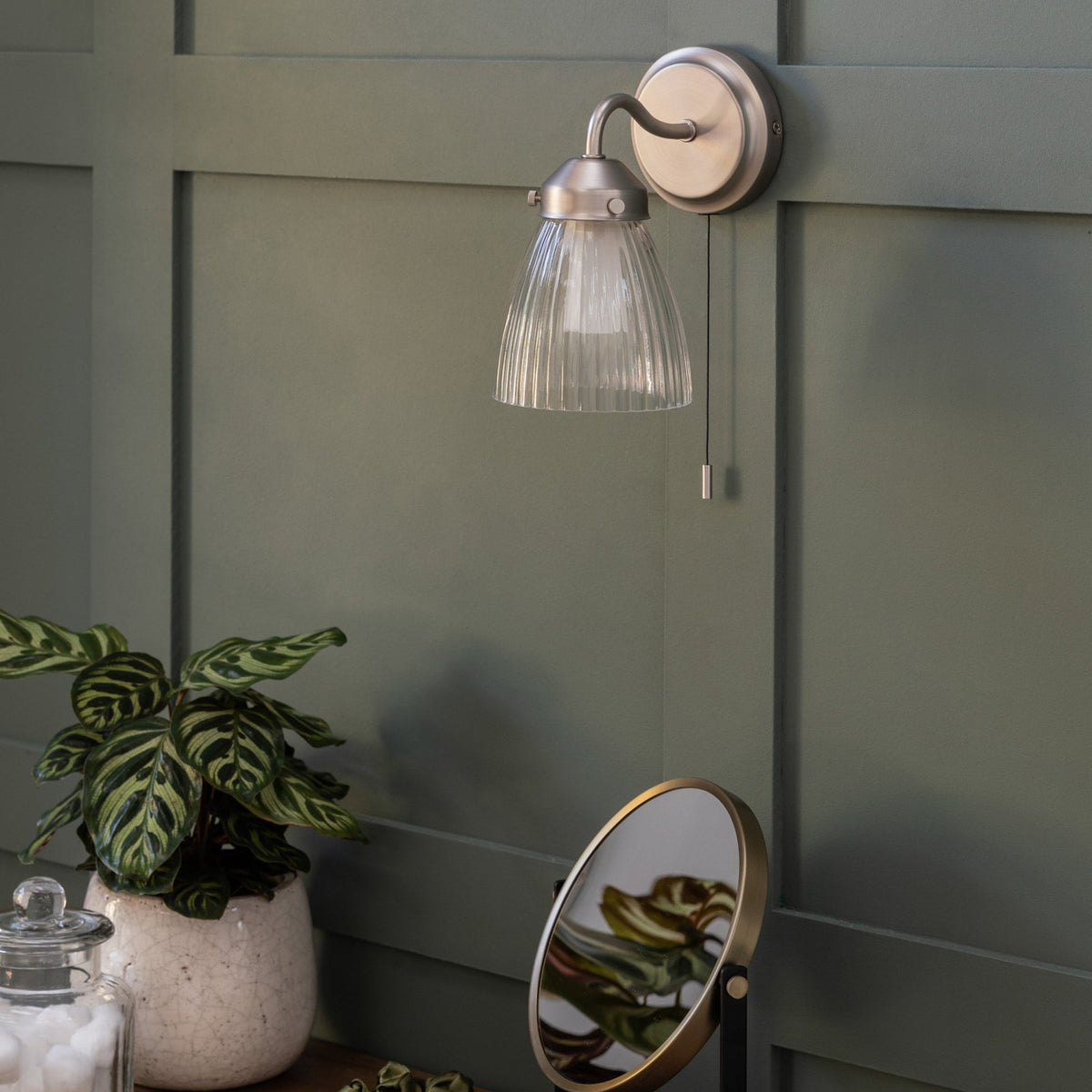 A wall-mounted light fixture with a ribbed glass shade and a pull chain, against a green paneled wall.