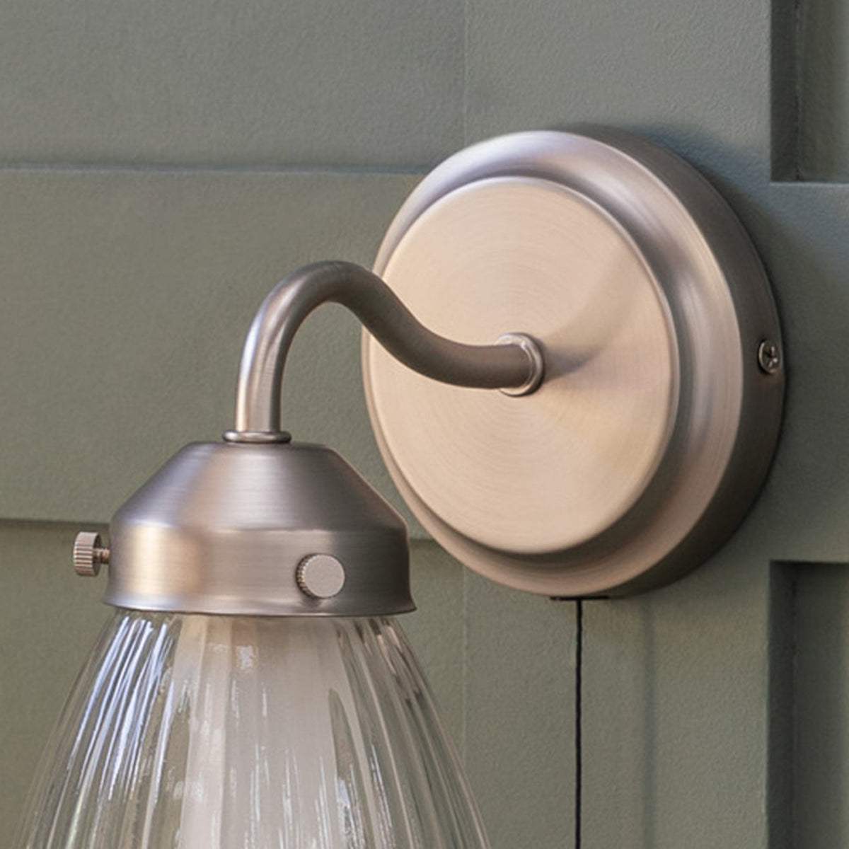 Brushed chrome wall light with a curved arm and a fluted glass shade, mounted on a green paneled wall.