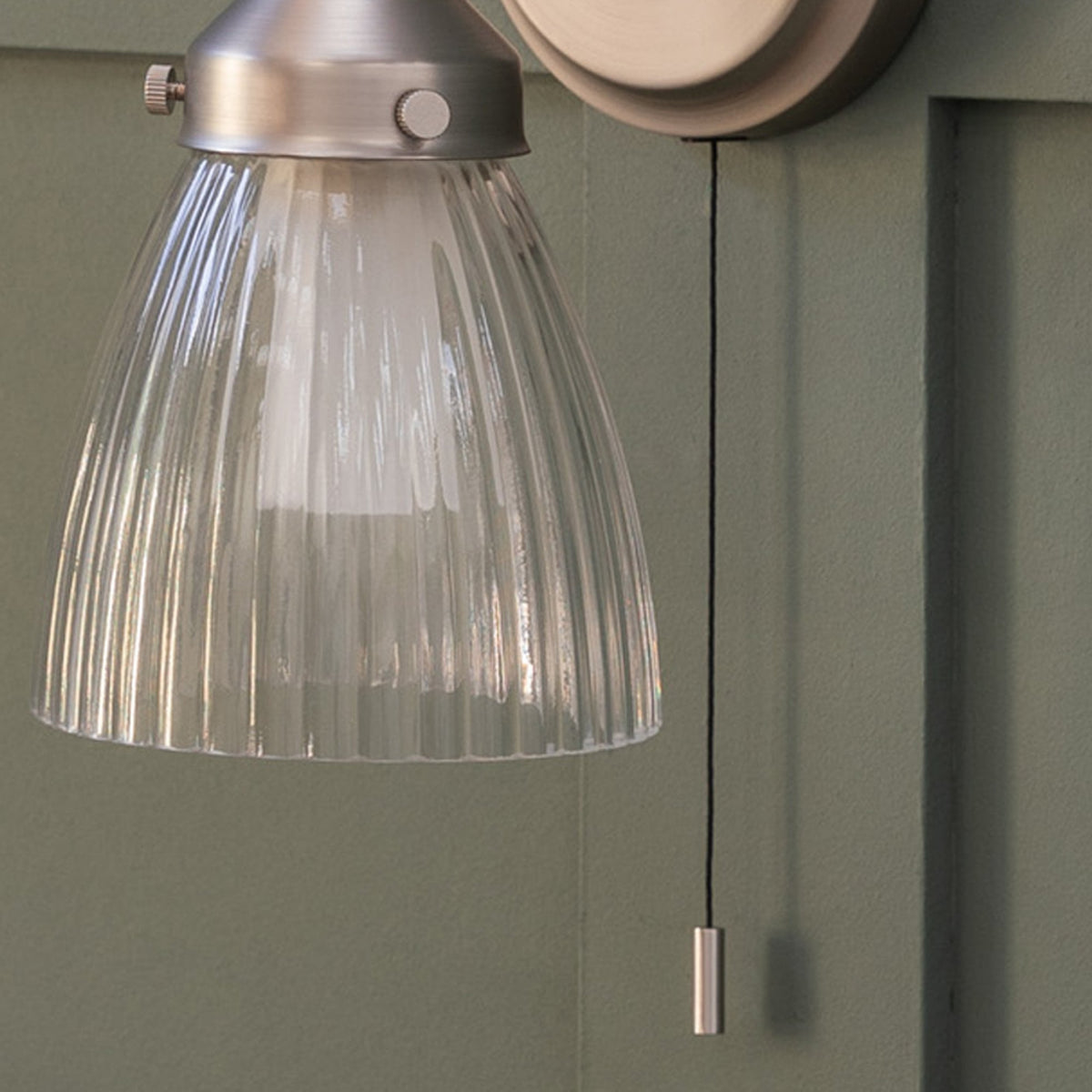 Close-up of a ribbed glass shade wall light with a brushed brass base and pull cord, against a green wall. 