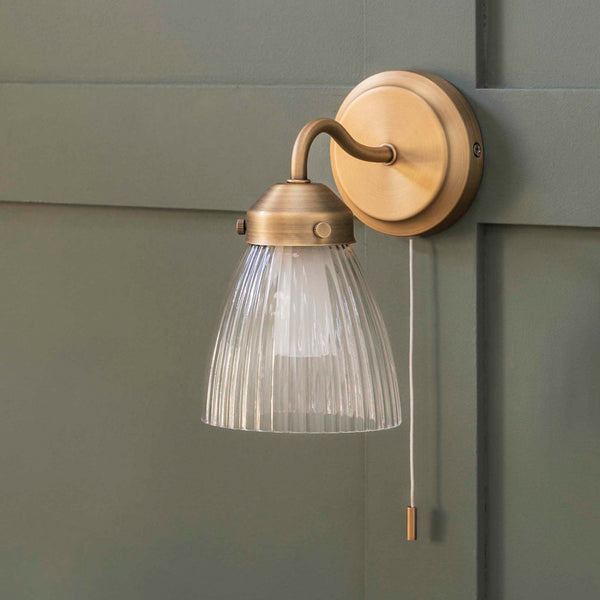 A brass wall light with a ribbed glass shade and a pull cord switch, mounted on a green paneled wall.