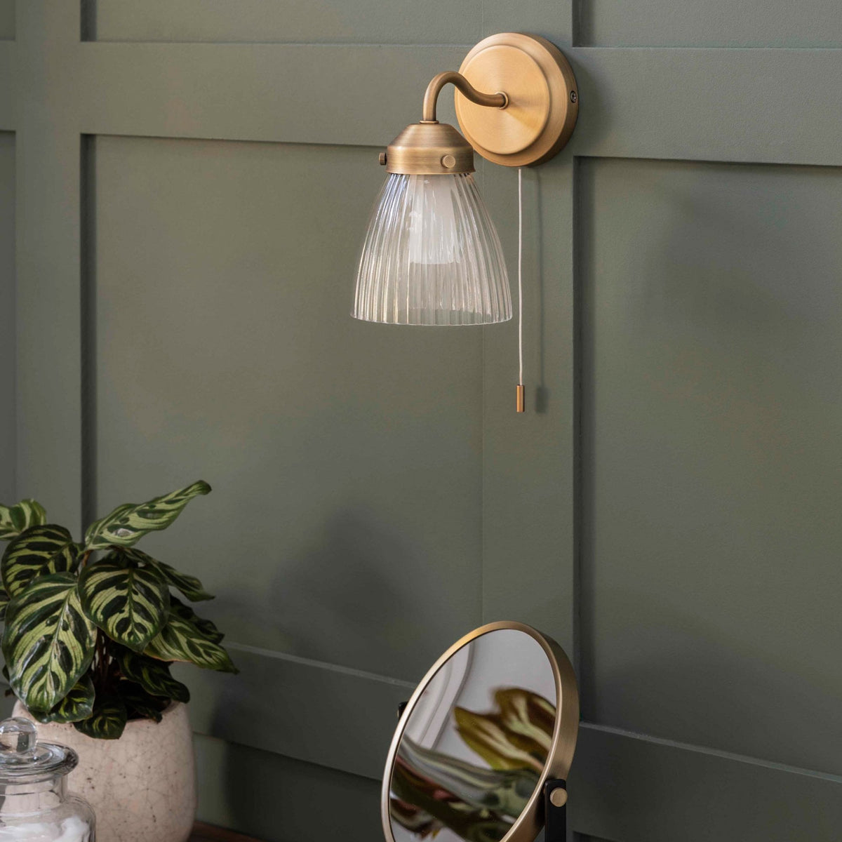 A brass wall light with a fluted glass shade, mounted on a dark green paneled wall. A plant and mirror are below.