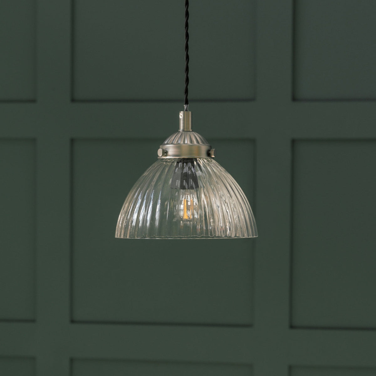A close up of a clear glass pendant light with a brass fitting and a visible filament bulb, against a dark green paneled wall.