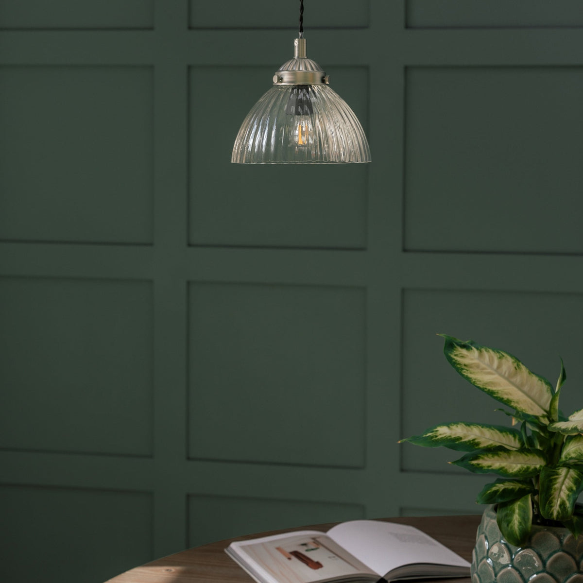 A ribbed glass pendant light with a warm bulb hangs above a wooden table with an open book and a plant.