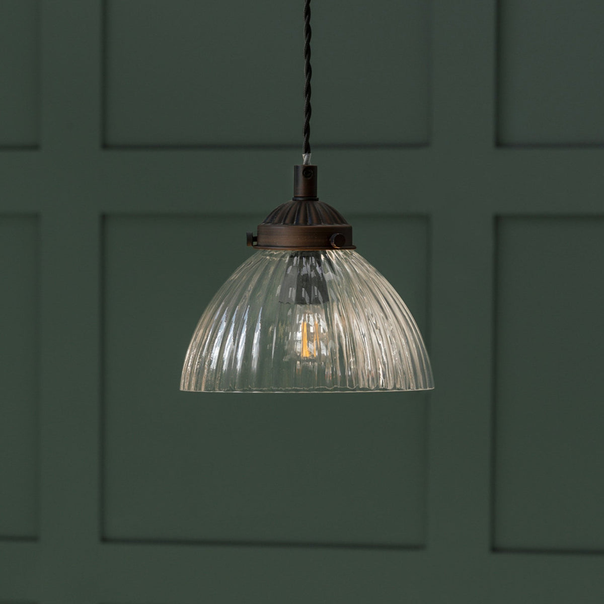 Ribbed glass pendant light with a bronze fitting and braided cord against a dark green paneled wall.