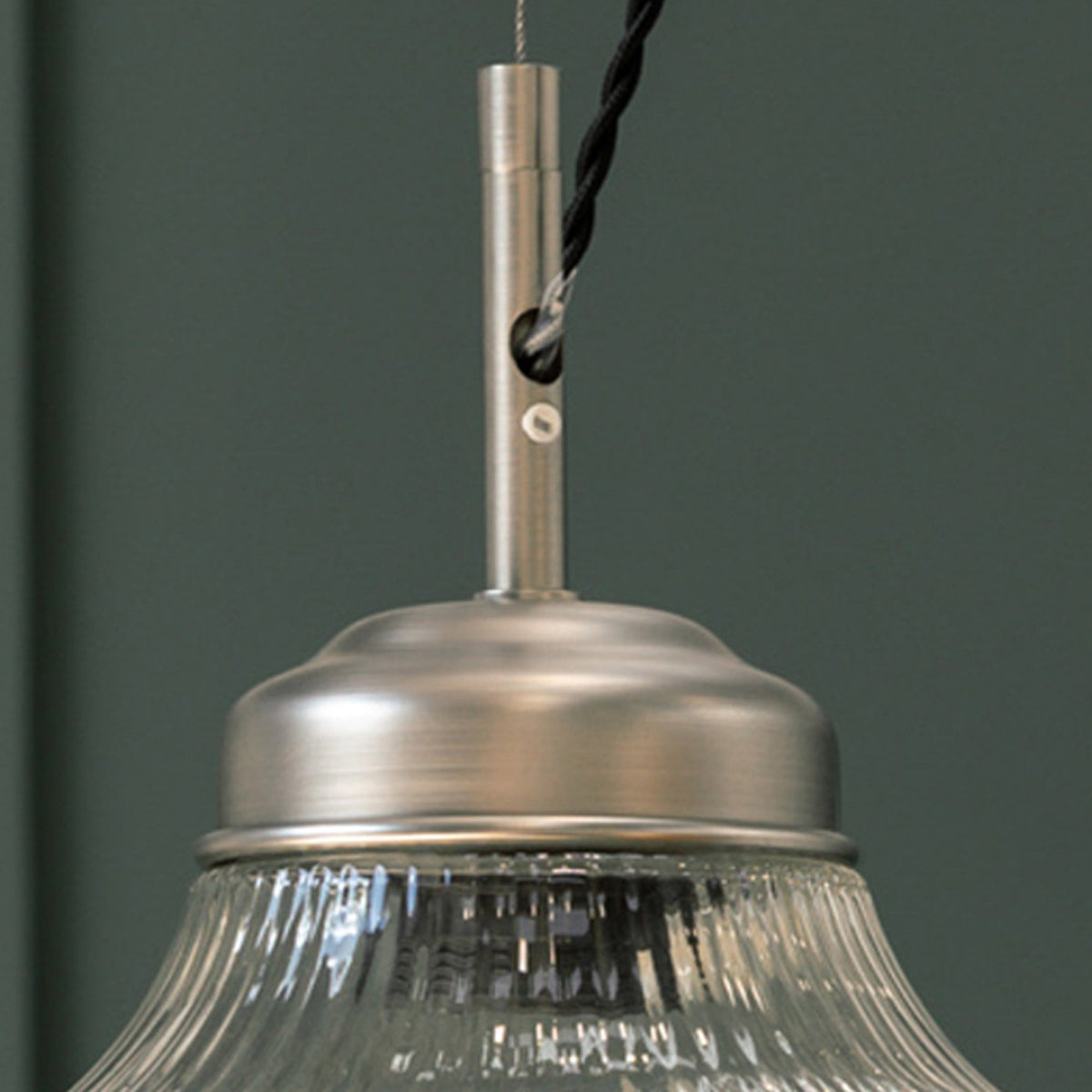 Close-up of a brushed metal pendant light fixture with a ribbed glass shade, against a dark green wall.