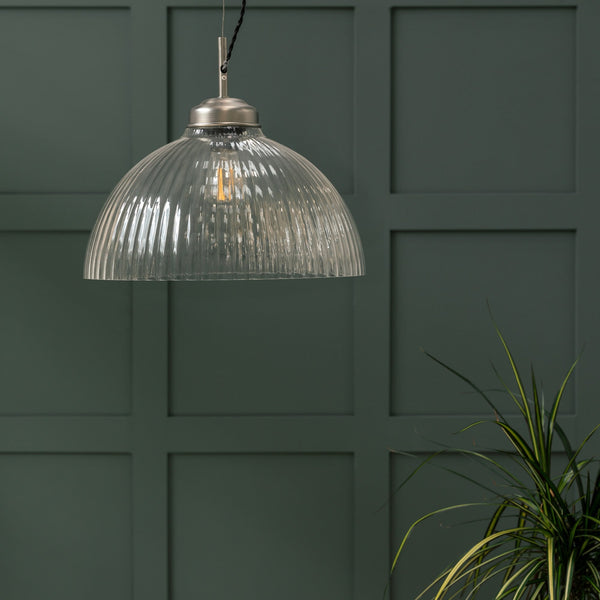 A ribbed glass pendant light with a brushed metal fitting hangs against a dark green paneled wall. A plant is in the bottom right corner.