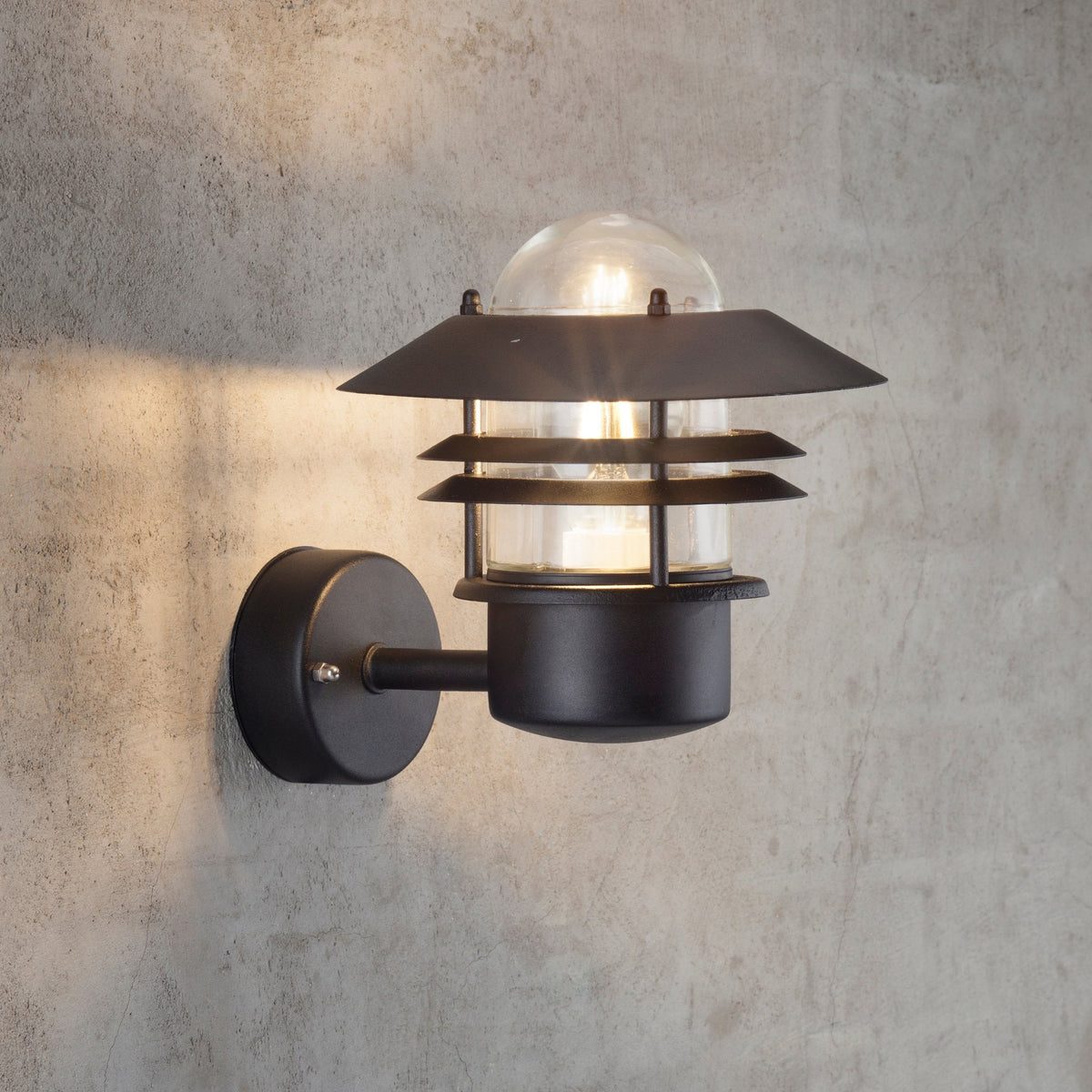 Black outdoor wall light with a clear glass shade illuminating a concrete wall.
