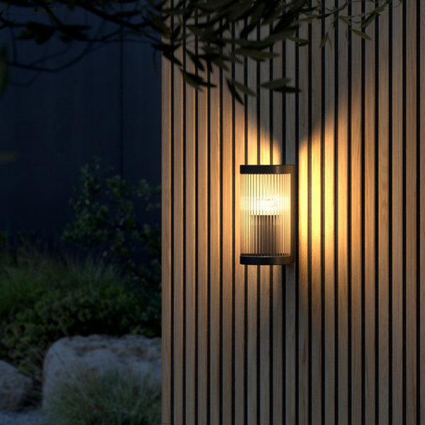 An outdoor wall light with a black frame and reeded glass casts a warm glow on a wooden wall.