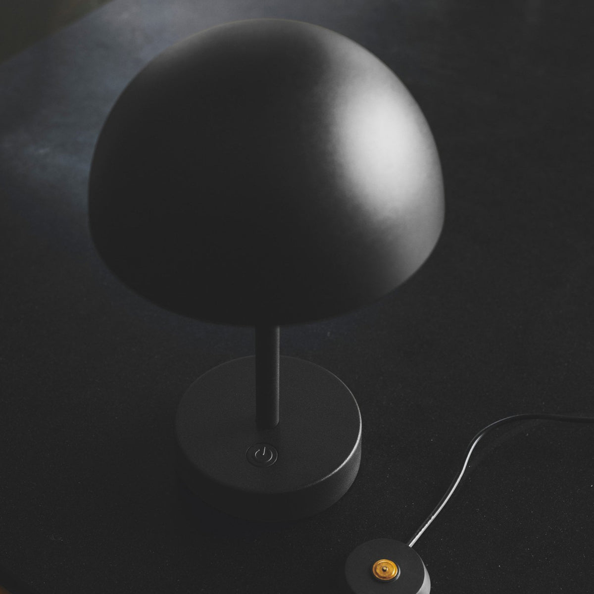 A black portable lamp on a dark surface with its charging cable and puck.