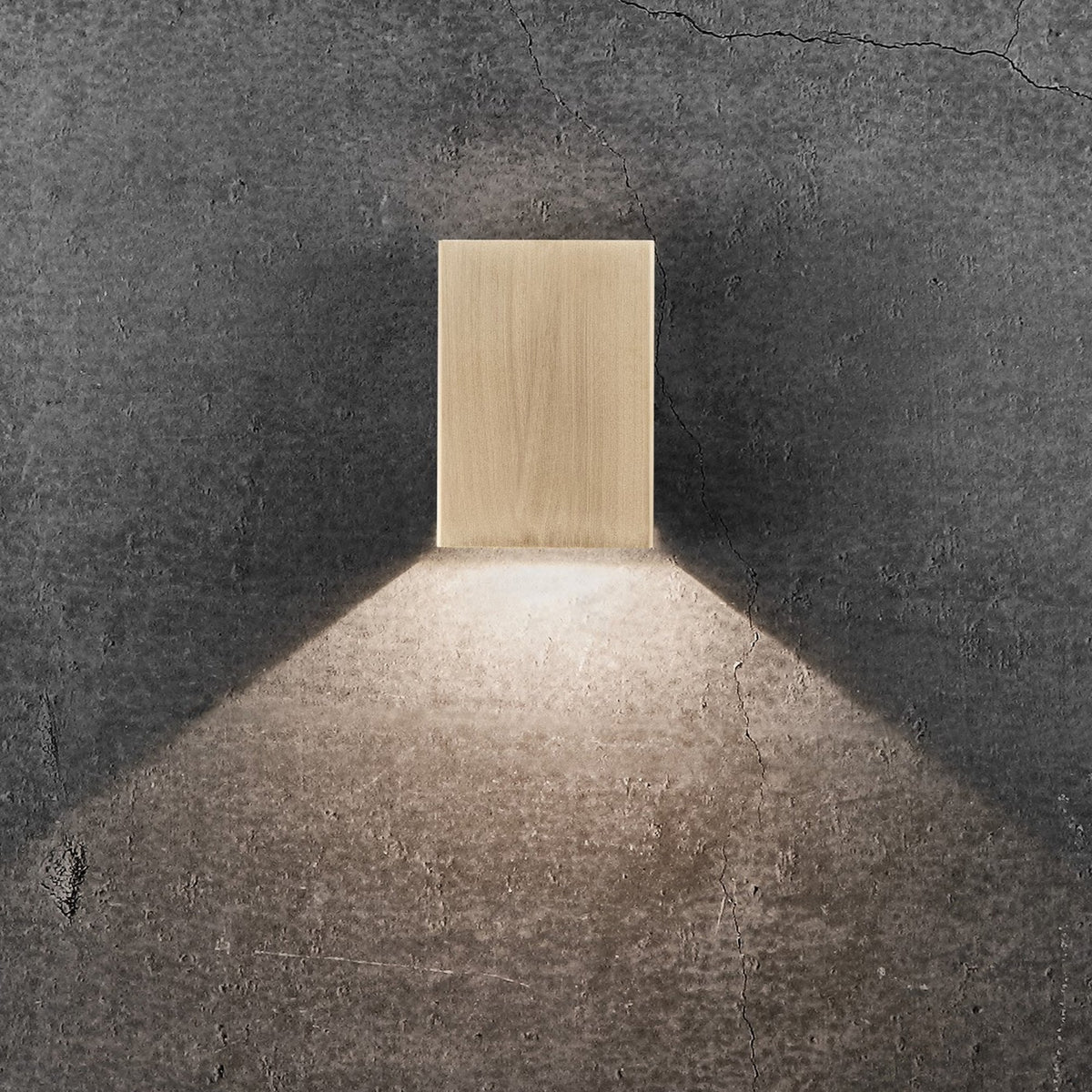 A golden rectangular wall light illuminating a textured grey wall, with light directed solely downwards.