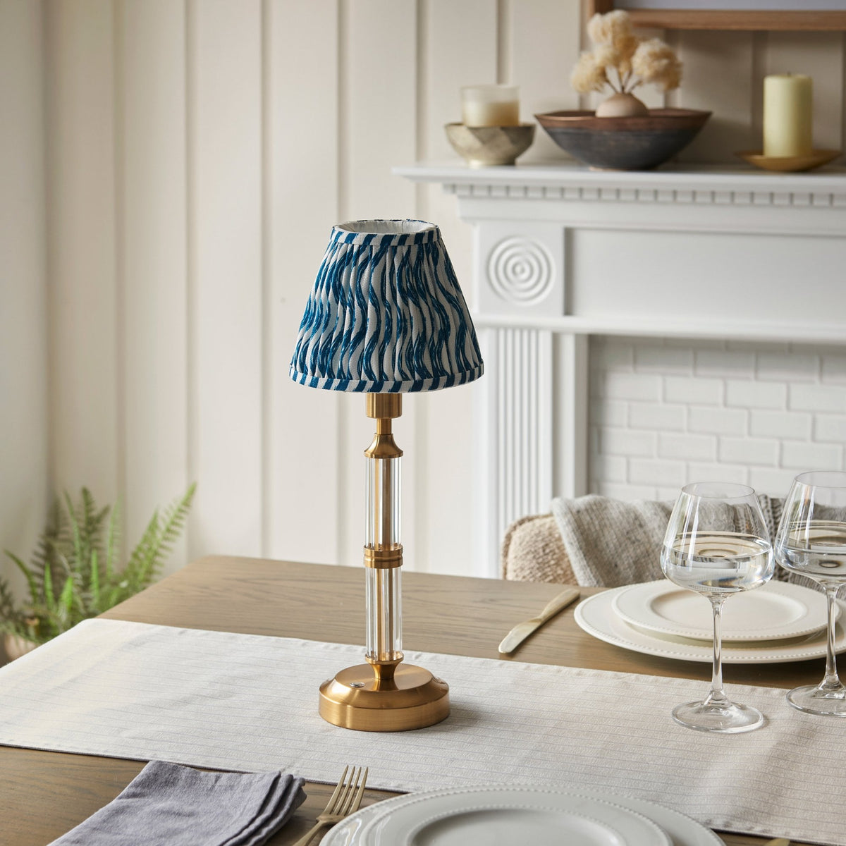 A small table lamp with a patterned shade sits on a dining table with place settings.