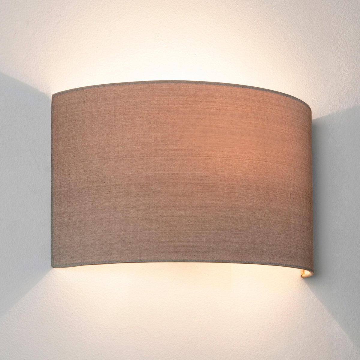 Wall light with a curved, rectangular, light brown fabric shade, casting light upwards and downwards on a white wall