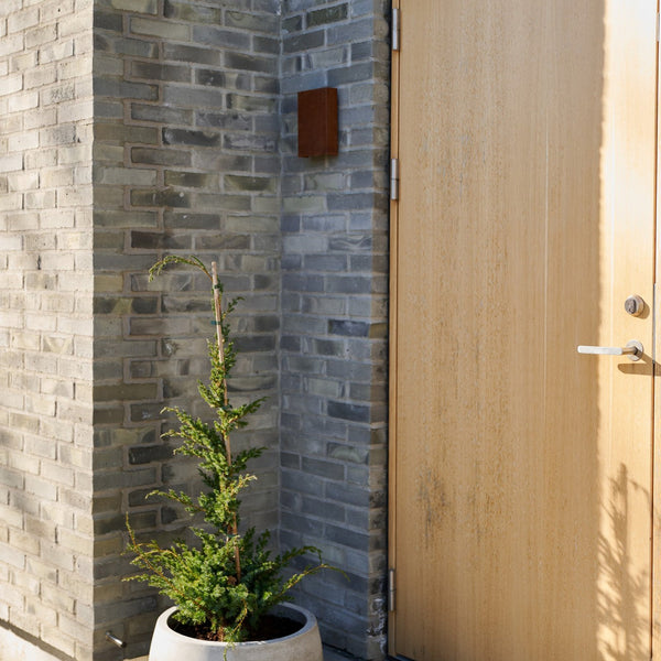 An outdoor wall light with a rusted finish on a grey brick wall next to a wooden door.