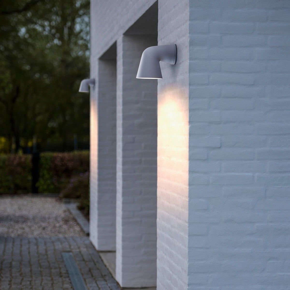 Two grey wall lights illuminate the exterior of a white brick building at dusk.