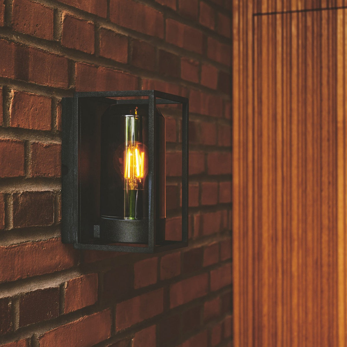A black outdoor wall light with a glowing filament bulb mounted on a brick wall.