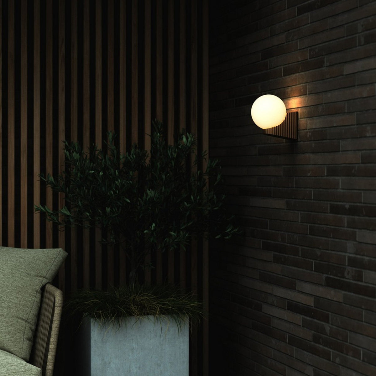 A round wall light illuminates a dark brick wall next to a potted plant and seating area.