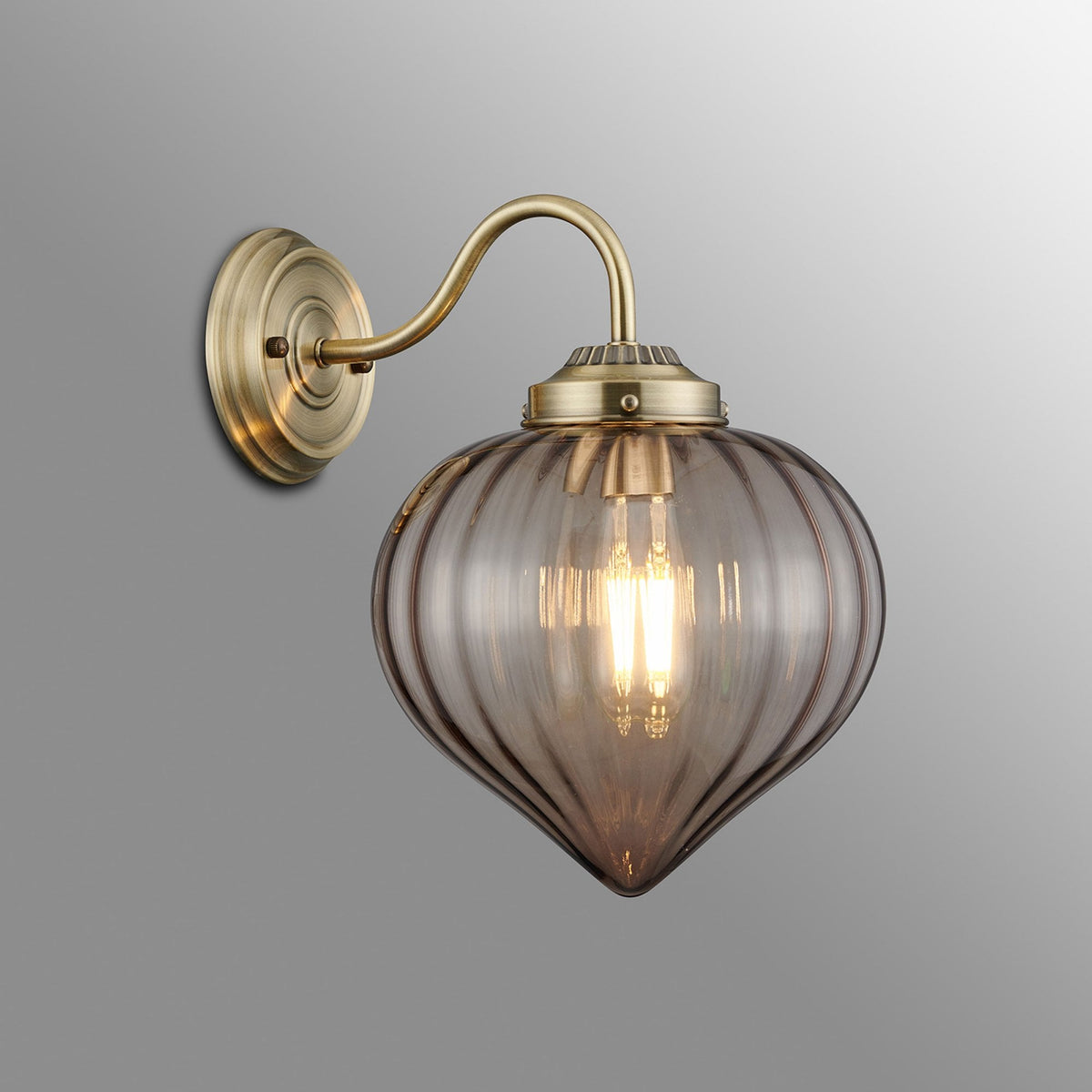 A brass wall light with a smoked, fluted glass shade housing a lit filament bulb.