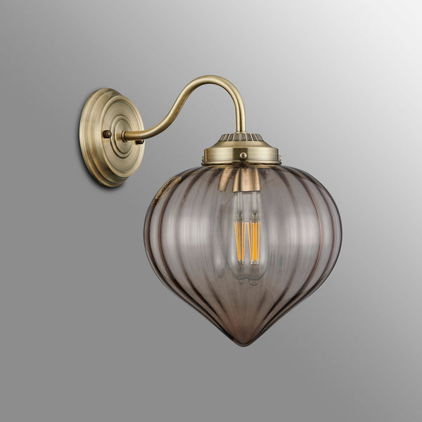 A brass wall light with a smoked, fluted glass shade housing an unlit filament bulb.