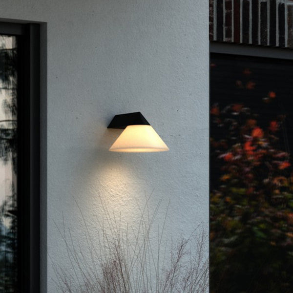 A modern outdoor wall light with a conical white shade illuminates a concrete wall at dusk.