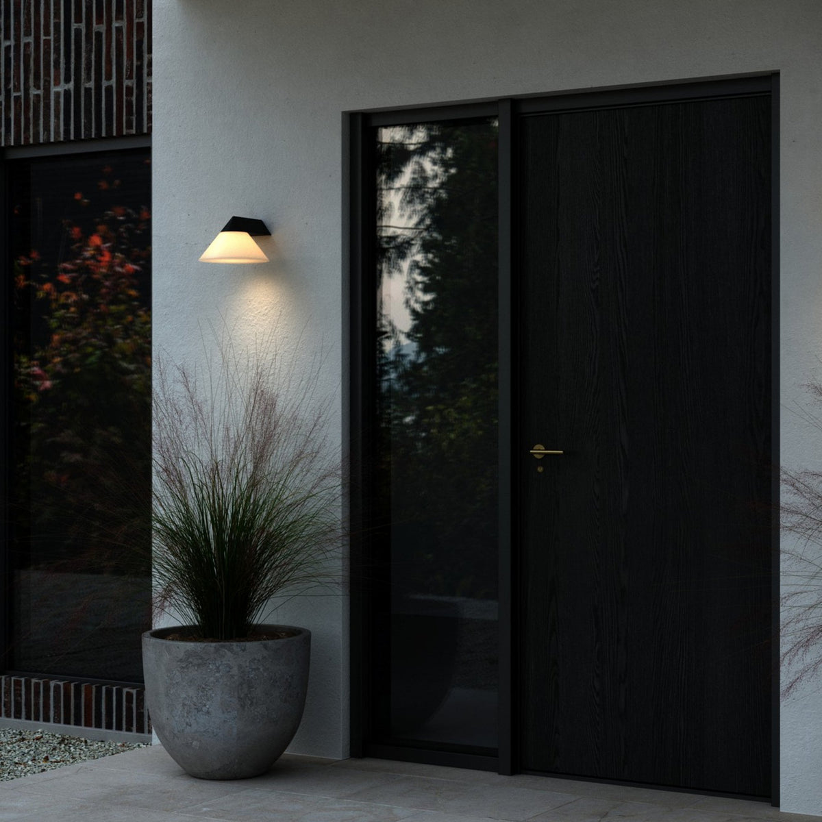 An illuminated outdoor wall light next to a dark modern door and potted plant.