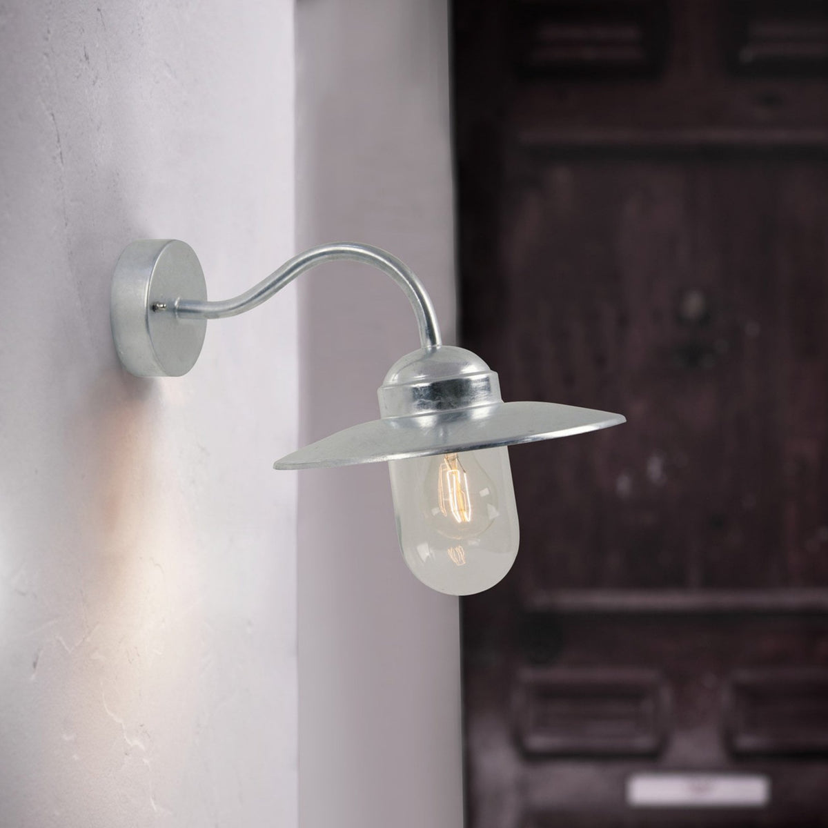 Silver outdoor wall light with a curved arm, a wide brim, and a clear glass bulb casing.