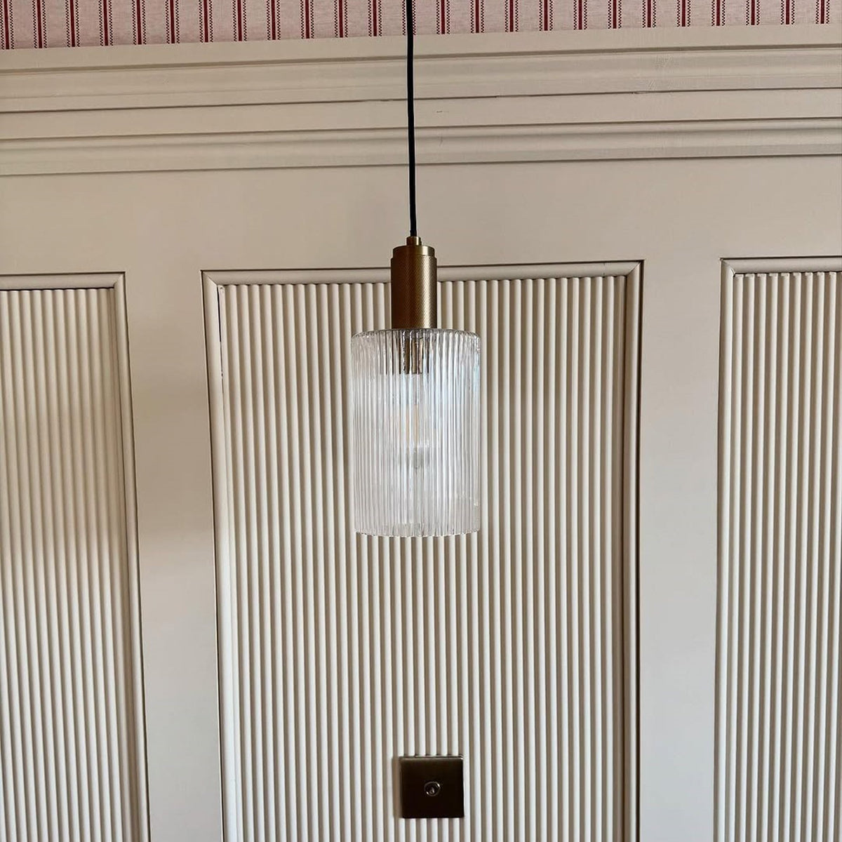 A fluted glass pendant light with a brass top hangs against a white fluted wall.