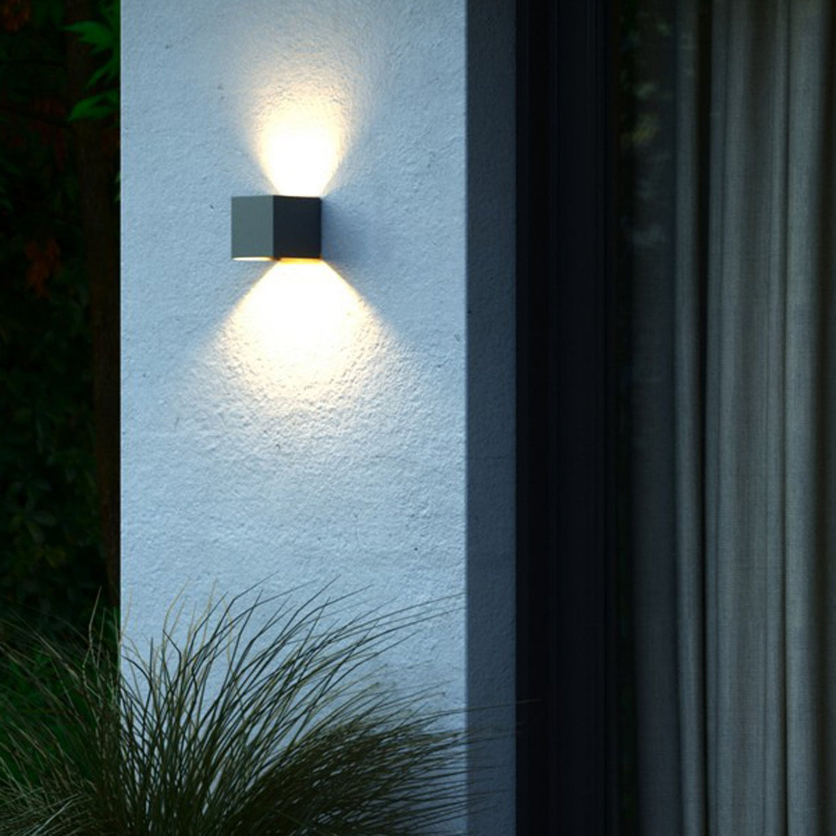 An anthracite cube-shaped outdoor wall light illuminates an exterior wall with warm light.