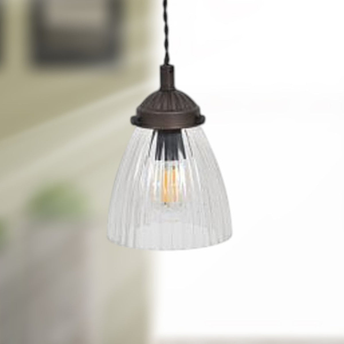 A close-up of a pendant light with a clear, ribbed glass shade and a dark bronze fixture. It hangs from a twisted cord.