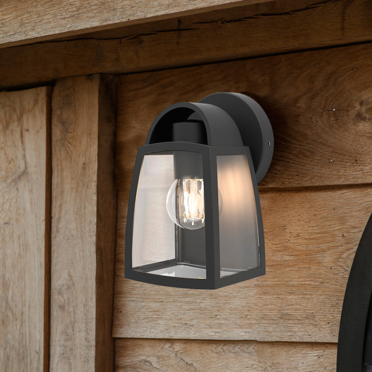 Black outdoor wall light with clear glass panels and an exposed filament bulb, mounted on rustic wooden siding.