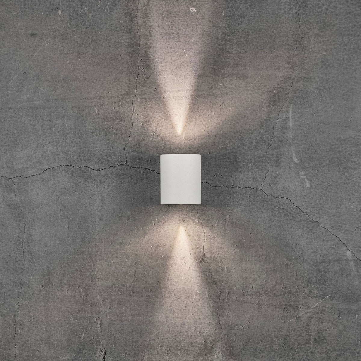 A white cylindrical outdoor wall light illuminates a grey concrete wall with two beams of light.