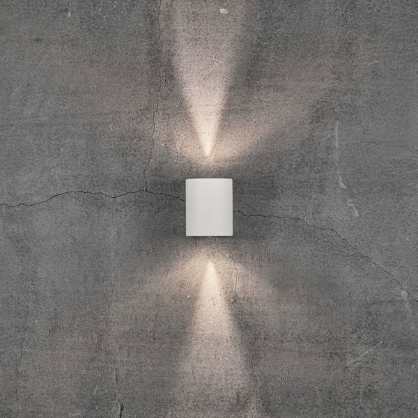 A white cylindrical outdoor wall light illuminates a grey concrete wall with two beams of light.