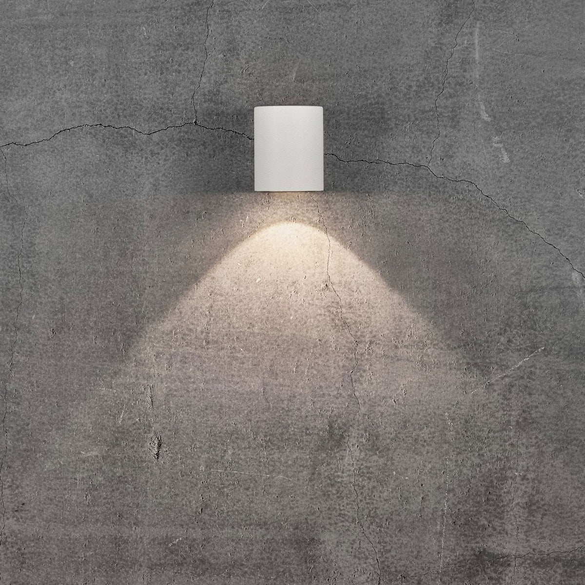 A white cylindrical outdoor wall light illuminates a grey concrete wall with a single beam of light.