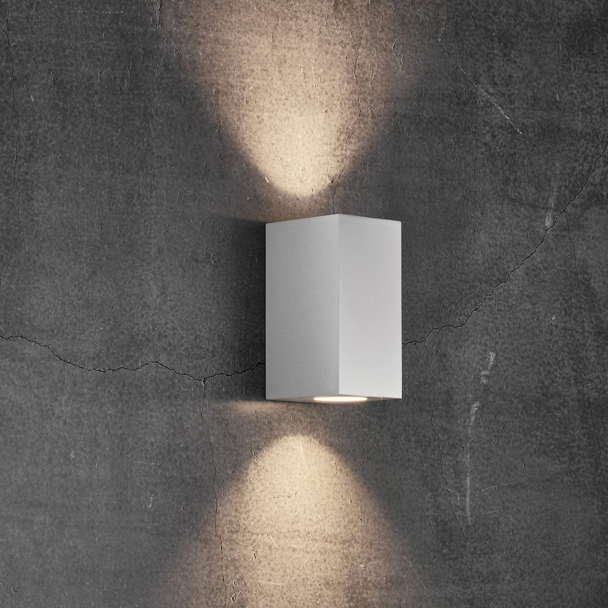 White rectangular outdoor wall light casting beams of light upwards and downwards on a dark wall.