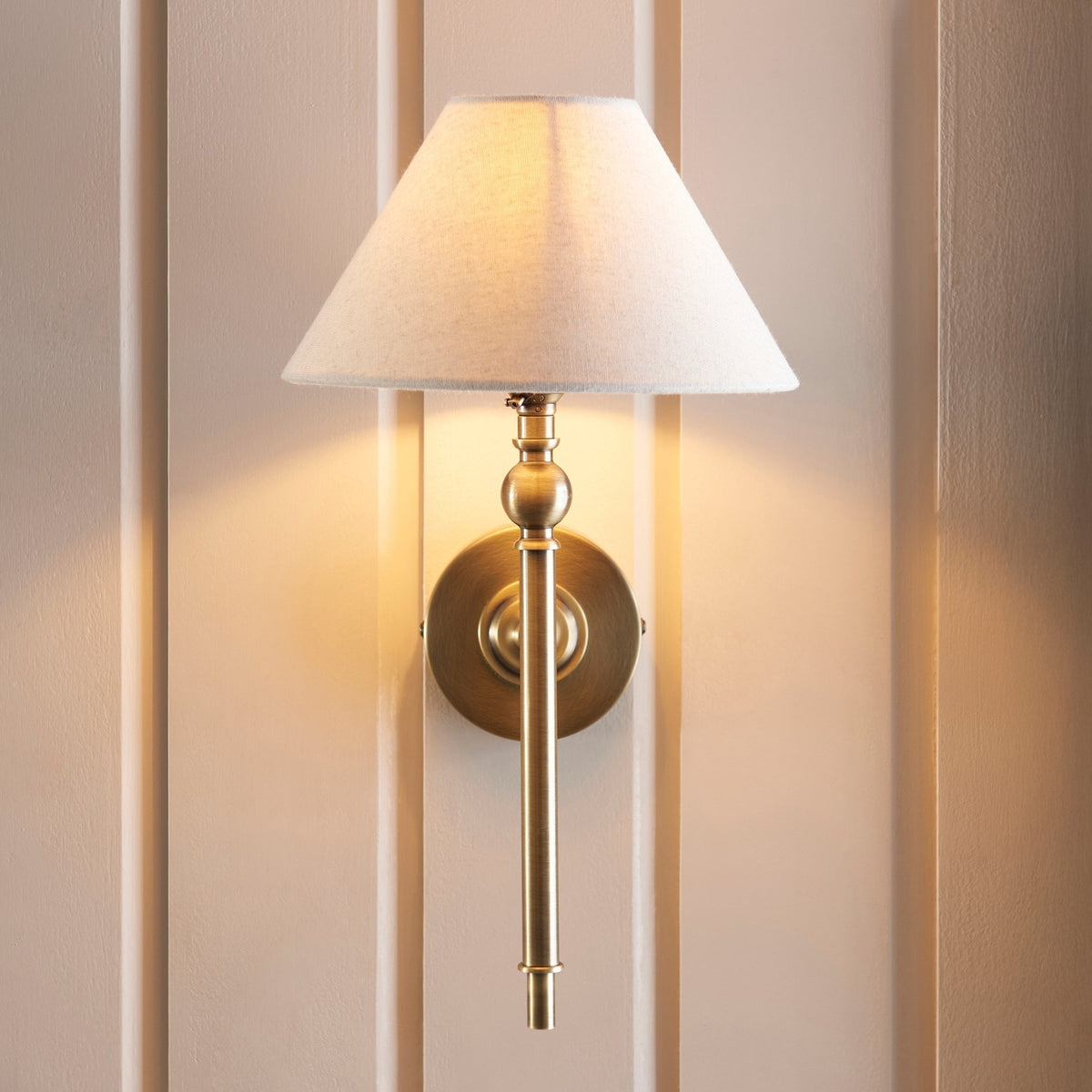 A brass wall light with a cream fabric shade illuminates a paneled wall.
