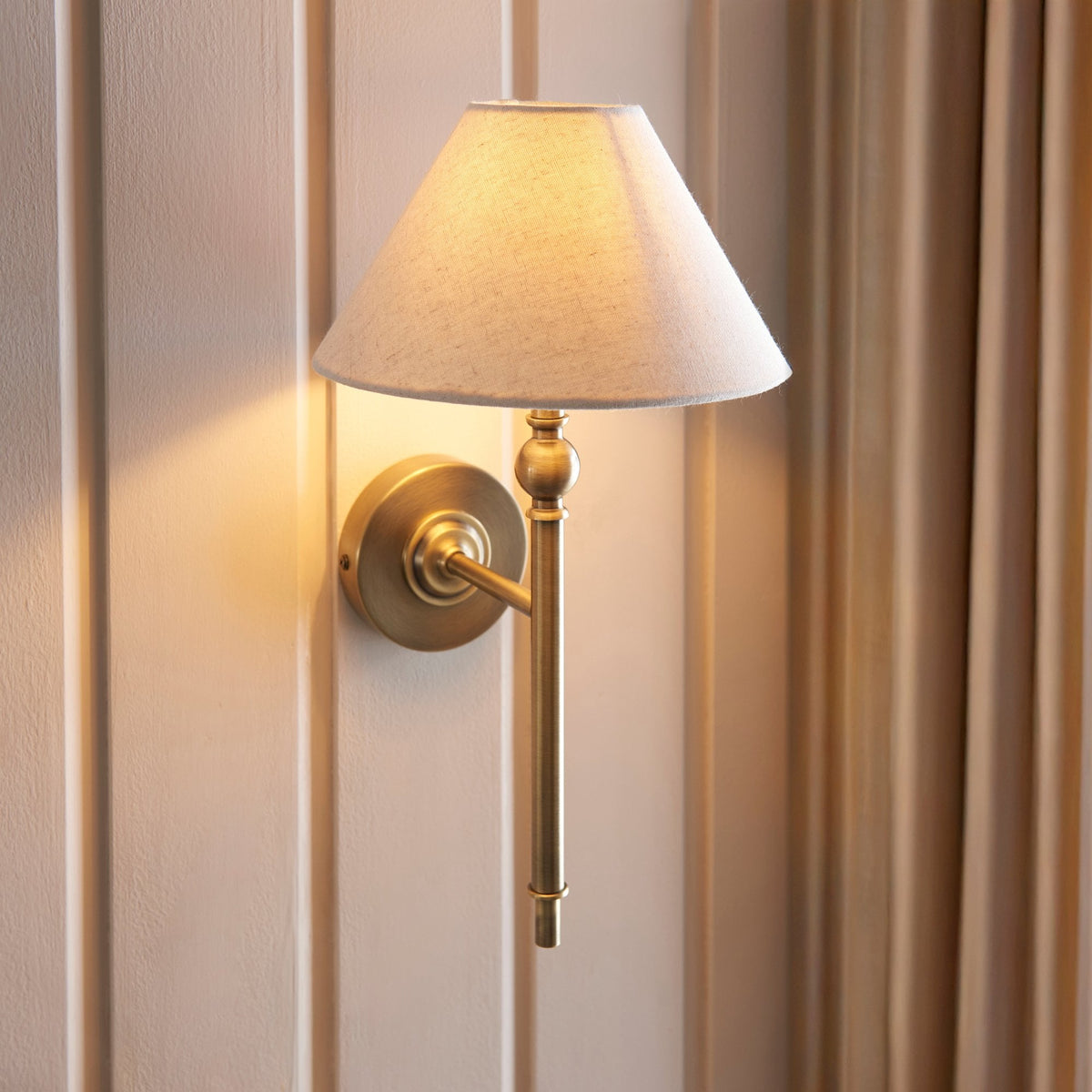 A brass wall light with a cream fabric shade casts a warm glow on a paneled wall.