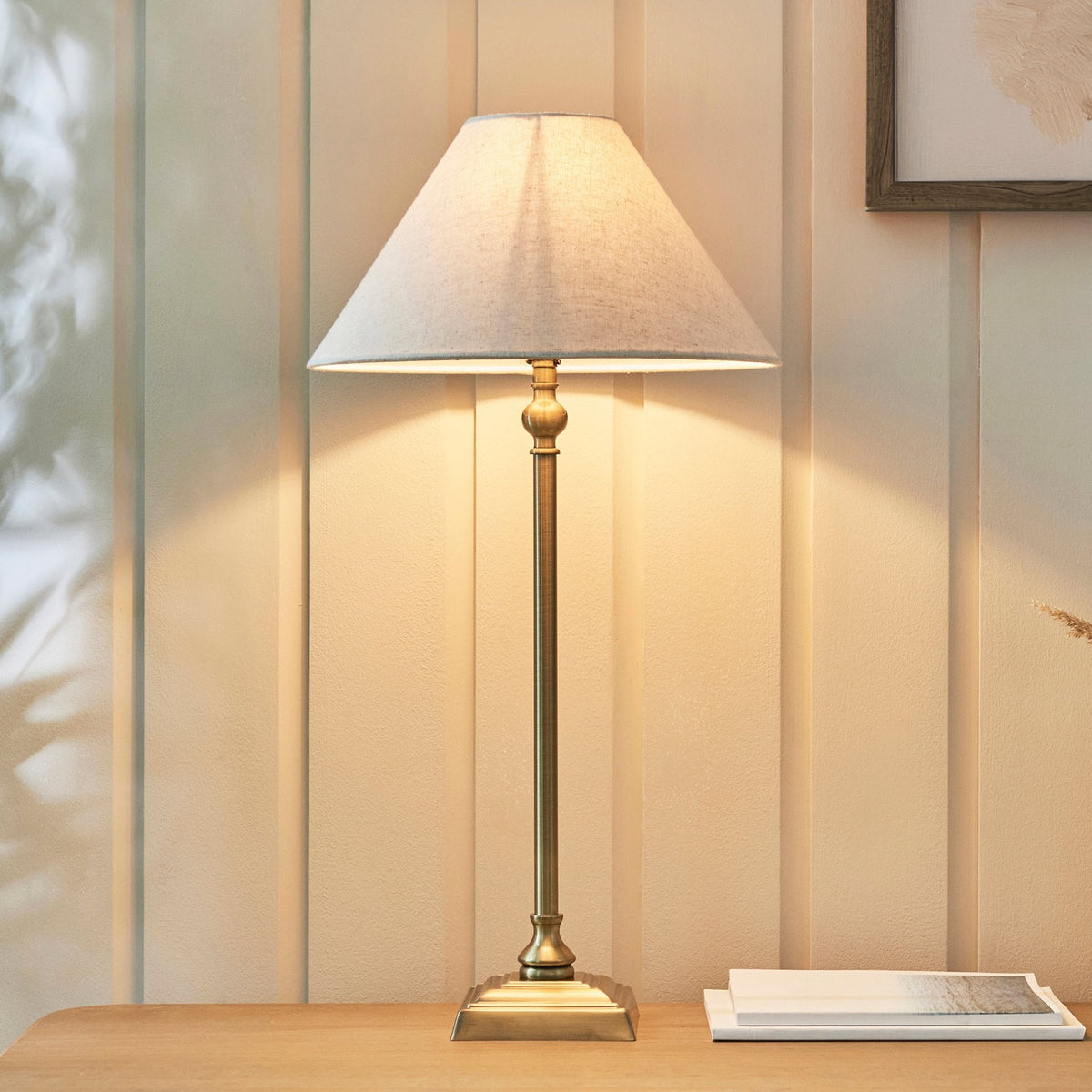 A brass table lamp with a light linen shade illuminates a wooden surface next to a framed picture.